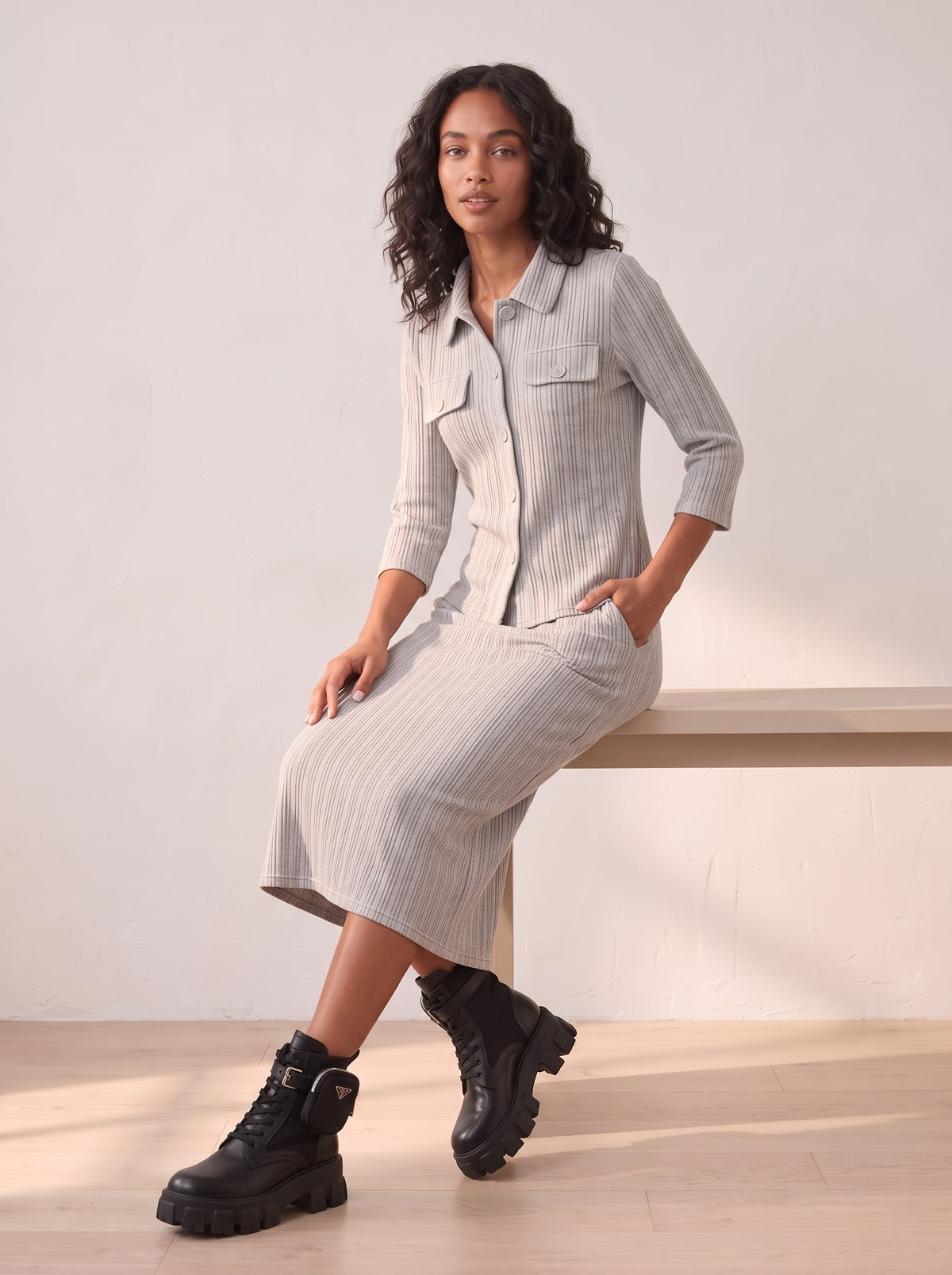 Seated model wearing Raya grey pleated shirt and matching straight midi skirt with chunky black ankle boots.